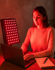 Person using a red light therapy device at a desk with a laptop and notebook.