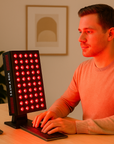 Person using a red light therapy device at a desk with a computer monitor.